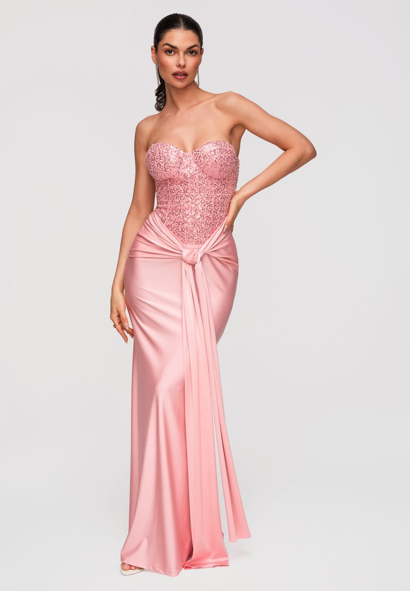 Femme portant une robe bustier rose à sequins avec une jupe en satin nouée en nœud à la taille, debout devant un fond uni.
