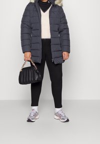 Cappotto imbottito grigio con cappuccio rifinito in pelliccia, leggings neri e sneakers argentate. Tiene una borsa nera trapuntata con dettagli a catena.