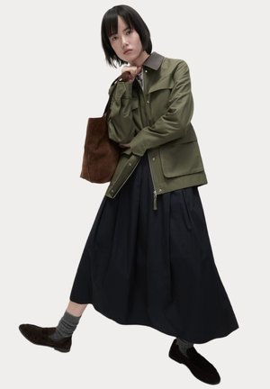 Chaqueta verde oliva con cuello marrón, de corte oversize. Combinada con una falda negra y un bolso de ante marrón. Calcetines a rayas grises y zapatos oscuros.