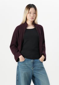 Ung kvinne med blondt hår iført en mørk lilla zip-up cardigan over en svart topp og blå jeans, med hendene i lommer, står foran en hvit bakgrunn.