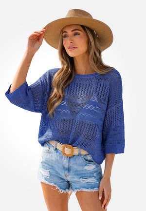 Femme portant un pull en tricot bleu, un short en jean déchiré avec une ceinture tissée, et un chapeau de paille à large bord touchant le bord avec une main.