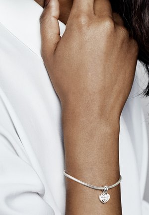 Silberarmband mit einer strukturierten Band und einem Herzanhänger, auf dem "Mama" eingraviert ist, getragen an einer Hand gegen ein weißes Hemd.