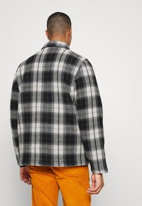 Herren Flanellhemd, schwarz-weißes Karomuster, weiche Textur, mit Kragen, gezeigt über orangen Cordhosen getragen.