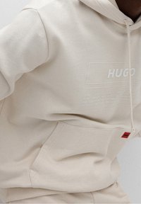Felpa beige realizzata in misto cotone, con tasca frontale, cappuccio regolabile con cordino e logo stampato con testo sul davanti. Dettaglio etichetta rossa.