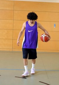 Lilla Nike tank top med hvide detaljer, sorte shorts og lyserøde sneakers. Holder en basketball, står på et gymgulv med vægge af træ.