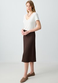 T-shirt blanc à col en V associé à une jupe midi froissée marron avec un ourlet en forme de scallop. Porté avec des mocassins marron clair. Design simple.
