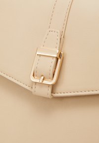 Beżowa skórzana torba z ozdobną złotą klamrą, gładką fakturą i przeszyciami wzdłuż krawędzi. Klasyczny fason z klapą.