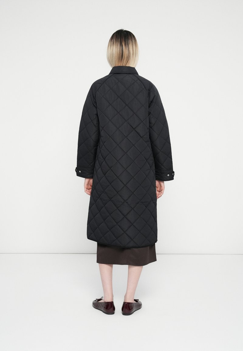 Lang zwart gewatteerd jasje met een diamanten patroon, met kraag, drukknopen op de manchetten, gedragen over een donkerbruine rok en bordeauxrode schoenen.