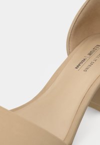 Gros plan sur une sandale à talon haut beige à bout ouvert avec une étiquette végan sur la semelle intérieure, sur fond blanc.