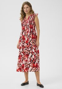 Robe maxi rouge à fleurs avec un bodice smocké, des manches à volants et une silhouette fluide ornée d'accents roses et blancs. Chaussures plates noires.