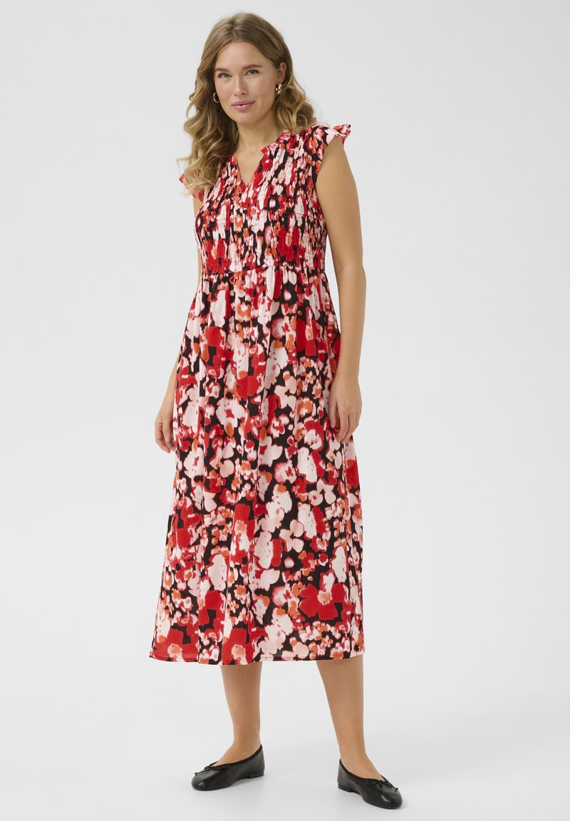 Robe maxi rouge à fleurs avec un bodice smocké, des manches à volants et une silhouette fluide ornée d'accents roses et blancs. Chaussures plates noires.