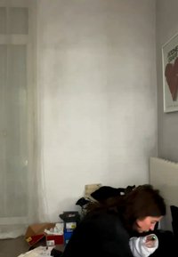 Osoba pochyla się w zagraconym pokoju z pudełkami i oprawionym plakatem z czerwonym sercem na ścianie.