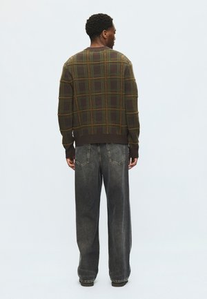 Ein Mann, der mit dem Rücken zur Kamera steht, trägt einen braun-grün karierten Pullover und locker sitzende, ausgebleichte schwarze Jeans vor einem einfarbigen Hintergrund.