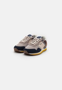 Zapatillas en beige, azul marino y marrón de ante con acentos blancos y suela amarilla. Presentan un collar acolchado y cordones para un ajuste adjustable.