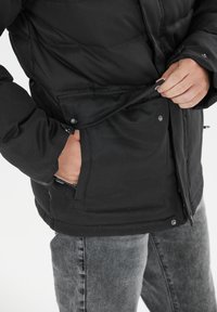 Veste en duvet noire avec fermeture à glissière et pression, équipée d'une poche latérale et d'un tissu texturé. Portée par-dessus un jean gris.