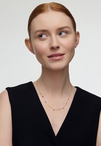 Złoty naszyjnik z małymi geometrycznymi zawieszkami i delikatnymi akcentami, noszony na ciemnej bluzce, na jasnym tle.