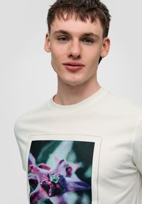 Lichtgekleurde katoenen t-shirt met een grote bloemenprint in paarse en blauwe tinten, voorzien van een ronde halslijn en korte mouwen.