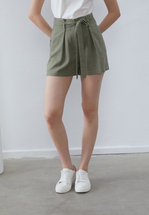 Olive green shorts with a tie belt, high waist, pleated front, and a tailored design, paired with white sneakers. Smooth fabric texture.