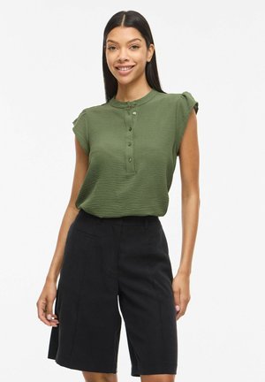 Femme aux longs cheveux noirs portant un chemisier vert texturé sans manches boutonné et un short noir à jambes larges, souriant à la caméra.