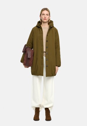 Kobieta stoi ubrana w oliwkowo-zielony płaszcz z kapturem, beżowy sweter, białe spodnie o szerokich nogawkach, brązowe buty i trzyma dużą brązową torbę w lewej ręce.