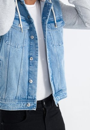 Persoon draagt een lichtblauw denimvest met metalen knopen over een wit overhemd en zwarte broek, en stelt de grijze capuchonkoorden van het vest af.