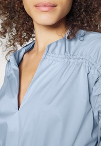 Chemise bleu clair, en tissu soyeux, avec un col en V et des détails plissés aux épaules. Texture lisse avec un tissu drapé, design minimaliste.