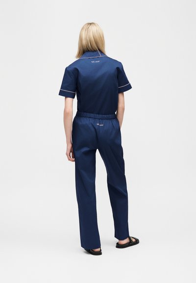 Ξανθιά γυναίκα όρθια, φορά μπλε ναυτικό κοντομάνικο πουκάμισο και ταιριαστό παντελόνι με ελαστική ζώνη μέσης και μαύρα σανδάλια, με την πλάτη προς τα εμπρός.