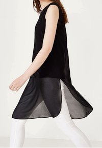 Black sleeveless tunic with a flowing design, featuring a translucent lower hem and a straight silhouette. Worn over white pants.