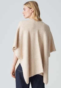 Poncho en tricot beige avec texture côtelée, manches larges et ourlet fendu. Matériau doux qui tombe délicatement sur le dos.