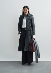 Trench-coat long en cuir gris foncé avec fermeture à boutons, porté sur une chemise blanche et un pantalon noir à jambes larges. Accessoires : un sac bordeaux et une écharpe grise.