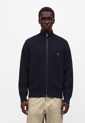 Homme portant un pull noir à fermeture éclair avec un petit logo sur la poitrine et un pantalon beige, debout devant un fond blanc uni.