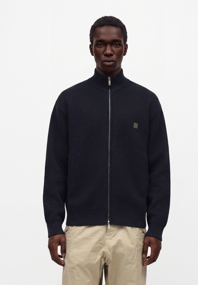 Homme portant un pull noir à fermeture éclair avec un petit logo sur la poitrine et un pantalon beige, debout devant un fond blanc uni.