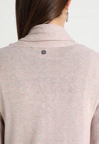 Žena s dlouhými hnědými vlasy, oblečená v světle růžovém pleteném svetru s malou černou značkovou nášivkou na horní části zad a stříbrnými kruhovými náušnicemi.