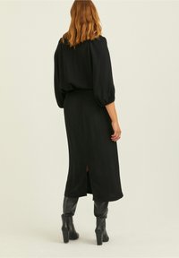 Schwarzes Midi-Kleid mit Puffärmeln, einer lockeren Passform und einem Schlitz im Rücken. Getragen mit knielangen schwarzen Stiefeln. Glatte Textur. Minimalistisches Design.