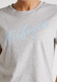 Camiseta de algodón gris con mangas cortas, que presenta un logotipo en escritura de color azul claro "Hilfiger" en el pecho. Textura suave y un ajuste casual.
