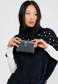 Billetera de teal acolchada con cierre a presión, detalles en plata, sostenida en manos contra un suéter oscuro con detalles de perlas.