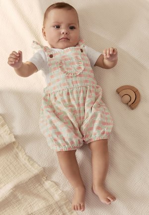 Beebi lamab kreemika teki peal, seljas pastelltoonis ruuduline kombinesoon ja valge särk, läheduses puidust vikerkaare mänguasi.