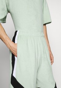 Personne portant un short en éponge vert clair et un haut à manches courtes assorti avec des bandes latérales noires et blanches, la main dans la poche.