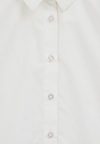 Biela bavlnená košeľa z prednej strany s štyrmi priehľadnými gombíkmi a špicatým golierom, pričom sú zobrazené detaily látky a šitia.