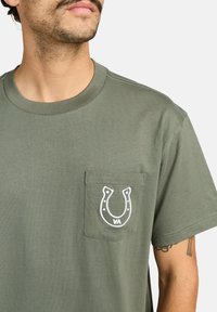 Camiseta de algodón color verde oliva con cuello redondo, que presenta un logo bordado de herradura blanca en el bolsillo del pecho izquierdo.
