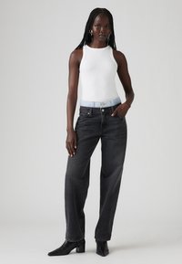 Fekete, magas derekú, bő szárú farmernadrág világoskék denim derékrésszel, fehér ujjatlan toppal és fekete, hegyes orrú bokacsizmával párosítva.