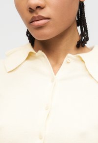 Gros plan sur une personne aux cheveux tressés portant un pull crème boutonné avec un large col.