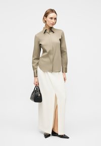 Kvinde iført en beige langærmet knappet skjorte, hvid slids maxi nederdel, sorte spidse flade sko og holder en lille sort håndtaske.