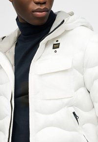 Κοντινό πλάνο ατόμου που φοράει λευκό μπουφάν από καπιτονέ ύφασμα με ετικέτα "Blauer U.S.A." και μπλε ζιβάγκο από μέσα.