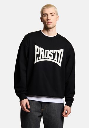 Młody mężczyzna o krótkich blond włosach, w czarnej bluzie z dużym białym napisem "PROSTO", białej koszulce pod spodem i ciemnych dżinsach.