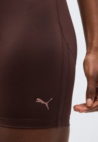 Κοντινό πλάνο ενός ατόμου που φορά καφέ σορτς Puma, δείχνοντας τη ραφή στο πλευρό και το λογότυπο Puma κοντά στον μηρό, με το δεξί χέρι χαλαρωμένο δίπλα.