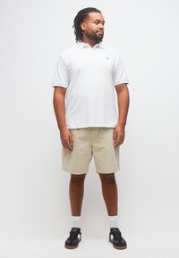 Άνδρας με πλεξούδες που φοράει λευκό πόλο μπλουζάκι, μπεζ σορτς, λευκές κάλτσες και μαύρα αθλητικά παπούτσια, στέκεται και κοιτάζει προς το πλάι.