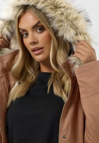 Ruskea huppu takki, jossa on tekoturkiksesta valmistettu reunus. Materiaali näyttää pehmeältä, ja siinä on vetoketjukiinnitys sekä lämmin vuori.