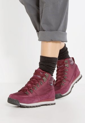 Persoon die opgerolde grijze broek draagt, donkere sokken en bordeauxrode wandelschoenen met metalen oogjes en gestructureerde zolen, staand op een witte ondergrond.