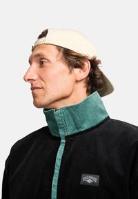 Black corduroy jacket with a teal ribbed collar and front panel, featuring a logo patch. Texture is prominent with vertical stripes.